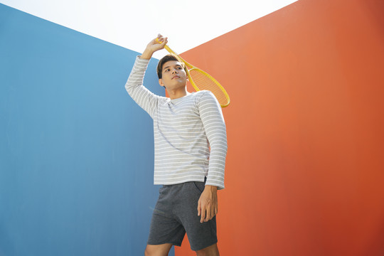 身着休闲服装、手持网球拍、背景为蓝橙色的迷人亚洲年轻男子。搞笑的姿势。打网球。举过头的姿势。