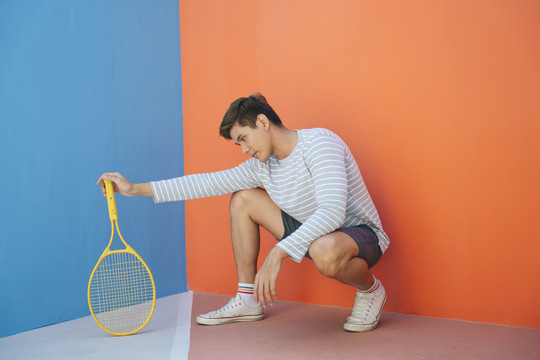 身着休闲服装、手持网球拍、背景为蓝橙色的迷人亚洲年轻男子。搞笑的姿势。打网球。以拍的姿势坐着。