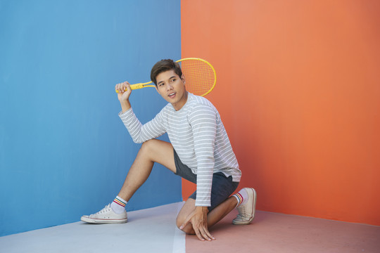 身着休闲服装、手持网球拍、背景为蓝橙色的迷人亚洲年轻男子。搞笑的姿势。打网球。拍拍姿势很酷。