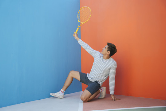 身着休闲服装、手持网球拍、背景为蓝橙色的迷人亚洲年轻男子。搞笑的姿势。打网球。这是我拍球拍的姿势。
