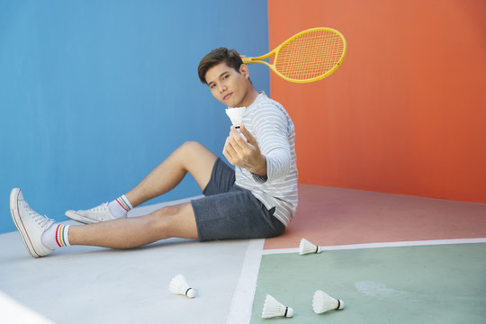递给一些亚洲网球运动员，他拿着球拍和羽毛球坐在地板上，背景是五颜六色的蓝色和橙色。