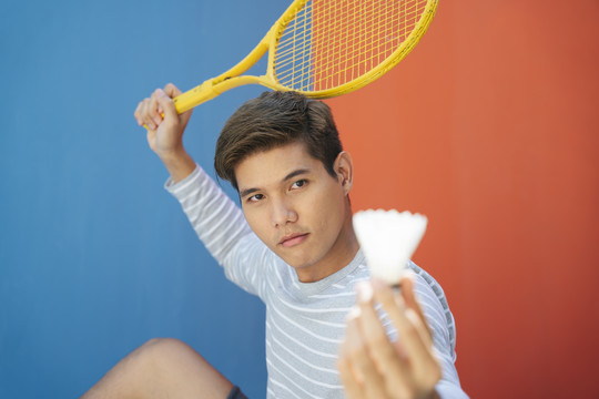 身着休闲服装、手持网球拍、背景为蓝橙色的迷人亚洲年轻男子。搞笑的姿势。打网球。我要打羽毛球。