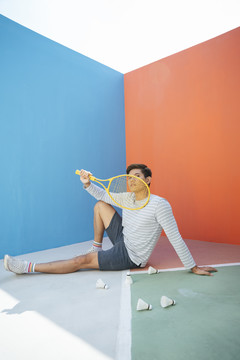 英俊的网球运动员亚洲男子坐在地板上，在五颜六色的蓝色和橙色背景上踢毽子。