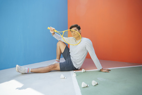 英俊的网球运动员亚洲男子坐在地板上，在五颜六色的蓝色和橙色背景上踢毽子。