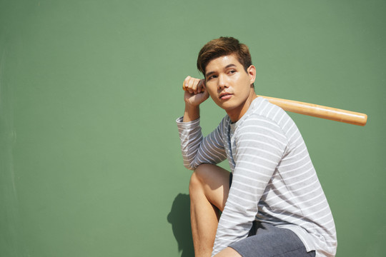 年轻英俊的亚洲男子拿着棒球棒在绿色背景上-复制空间。