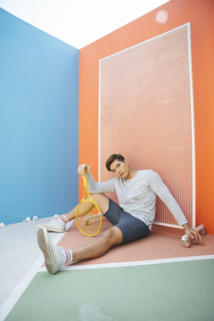 拿着黄色夹克的亚洲男子网球运动员坐在橙色背景的地板上。