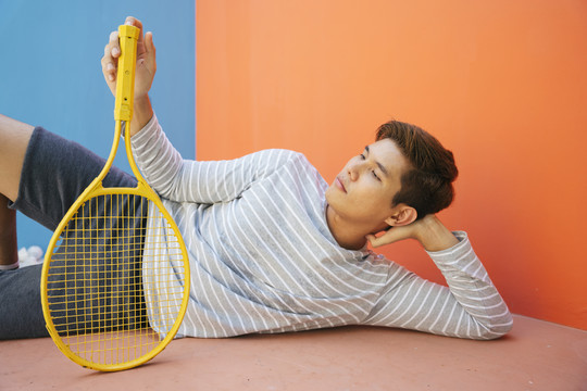 亚洲网球运动员躺在地板上用网球拍在橙色背景上。