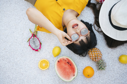 俯视图-戴着太阳镜、穿着黄色衬衫、躺在地上吃着热带水果的亚洲年轻女子。背景。
