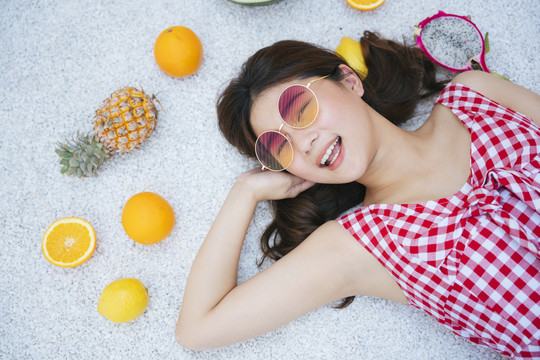 可爱美丽的亚洲年轻女子戴着时尚的眼镜，穿着红色的连衣裙躺在地上，上面放着热带水果。暑假时间。