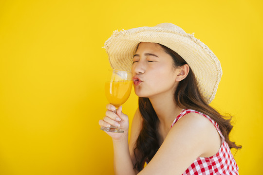 特写美丽的亚洲年轻女子戴着帽子拿着橙汁玻璃杯隔离在橙色或黄色的背景。