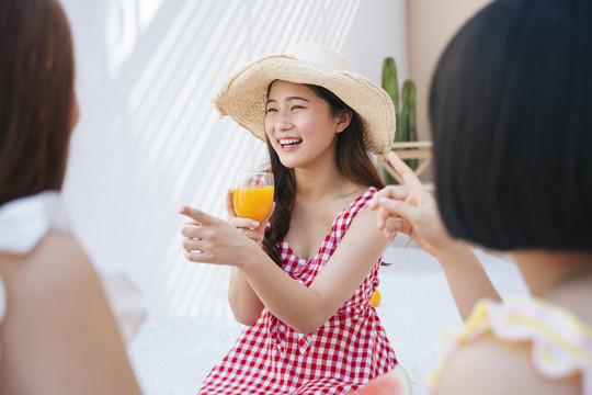 美丽的亚洲年轻女子戴着帽子，穿着红裙子，和朋友在夏日派对上喝着橙汁。