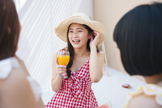 美丽的亚洲年轻女子戴着帽子，穿着红裙子，和朋友在夏日派对上喝着橙汁。