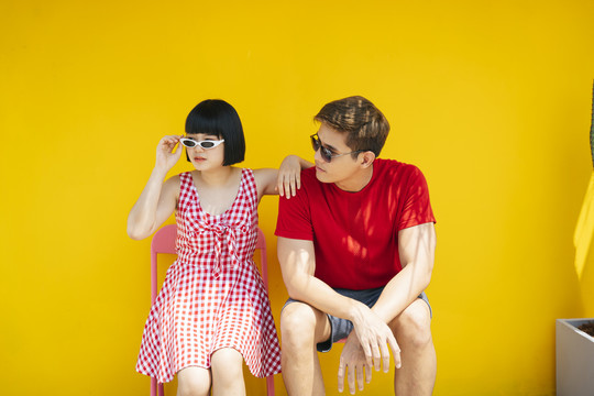 一对穿着红衬衫、戴着太阳镜的亚泰夫妇在橘黄色背景下坐在长凳上度过暑假。