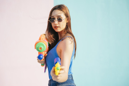 穿着蓝色背心的性感年轻女子手持水枪对准镜头。在柔和的粉蓝色背景上孤立。泼水节概念。