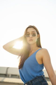 穿着蓝色背心衬衫，戴着太阳镜的性感美女肖像。性感的女人在日落时分摆出很酷的姿势。