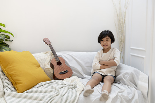 自信的亚洲男孩和四弦琴坐在客厅的沙发上。