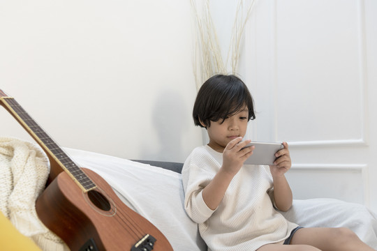 亚洲小男孩喜欢在智能手机上玩游戏，坐在起居室沙发上的四弦琴旁边。
