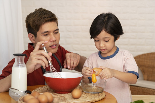 亚洲家庭烹饪活动-父亲看着儿子把蛋黄放进碗里。