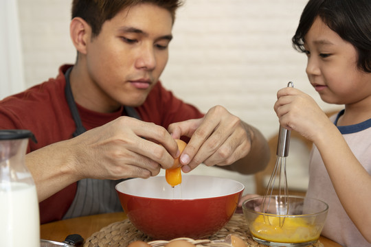 亚洲家庭烹饪活动-父亲将蛋黄从蛋壳中取出放入碗中，而儿子则在另一个碗中搅拌配料。