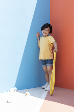 穿着黄色衬衫的可爱小男孩拿着网球拍，背景是五颜六色的蓝红色。地板上的羽毛球。对着墙摆姿势。