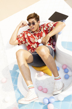 身着夏威夷衬衫、戴着墨镜躺在游泳池上的年轻英俊的亚泰男子的全长肖像。缤纷夏日时尚写真。