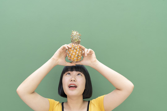 鲍勃发型泰国年轻女子举着菠萝在头上。隔离在绿色背景上。