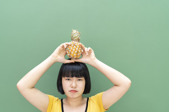 鲍勃发型泰国年轻女子举着菠萝在头上。隔离在绿色背景上。