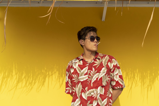 泰国帅哥穿着夏威夷红衬衫，戴着太阳镜，舔着黄色背景下的棒棒糖。夏季概念。
