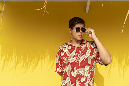 泰国帅哥穿着夏威夷红衬衫，戴着太阳镜，舔着黄色背景下的棒棒糖。夏季概念。