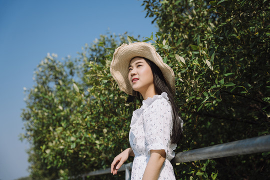 长发女子穿着连衣裙，戴着帽子，站在扶手旁，在蓝天下的树上微笑着。