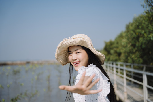 一个戴着帽子，穿着衣服的长发女人站在红树林旁的小码头上，把手伸向镜头。