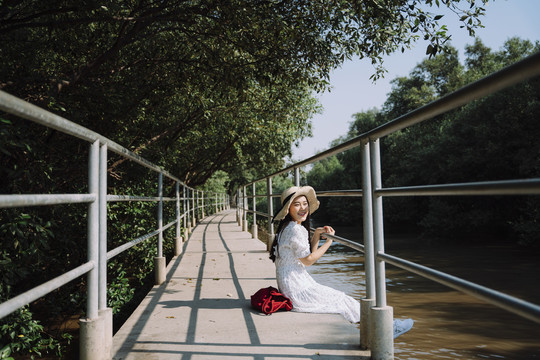 阳光明媚的日子里，一个戴着帽子的长发女人坐在红树林旁的小码头上，抓住码头的扶手。