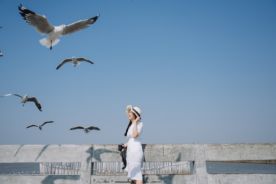 一位身穿白色连衣裙、头戴白帽的长发女子站在码头上，为蓝天上的海鸥拍照。