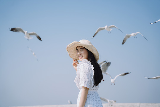长发白衣白帽女子与海鸥一起站在蓝天下的阳光下。