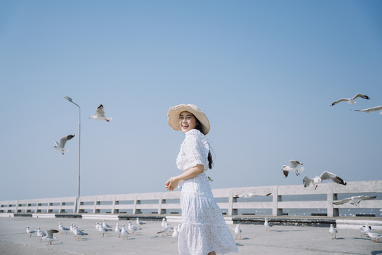 一个长发的女孩，穿着白裙子，戴着白帽子，在阳光下和站在码头上的海鸥玩耍。