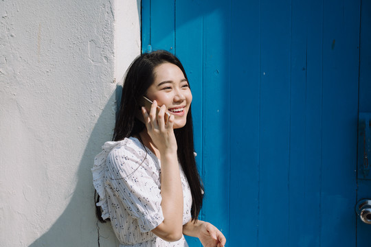 一位长发白衣女子站在蓝色的门口，拿着智能手机给她的朋友打电话。