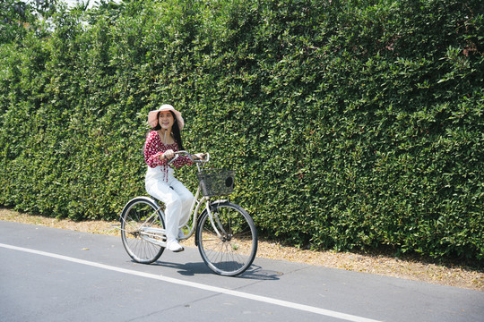 阳光明媚的日子里，穿着红衬衫的黑长发女孩和路边的大灌木丛一起骑自行车。
