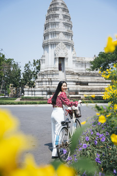 穿红衬衫的黑长发女孩在庙里走着，骑着自行车在黄花丛旁。