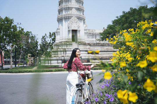 穿红衬衫的黑长发女孩在庙里走着，骑着自行车在黄花丛旁。