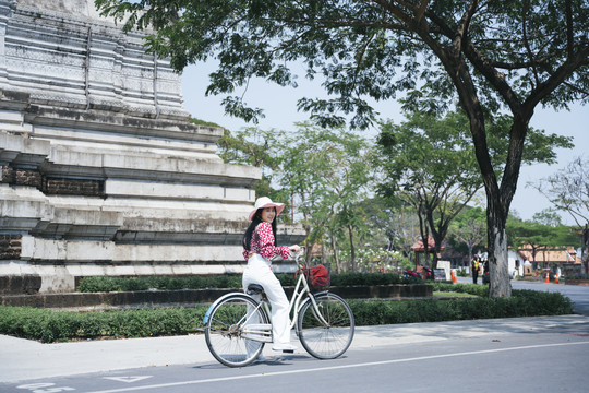 在阳光明媚的日子里，穿着红衬衫的黑长发女孩骑着自行车在寺庙里转。