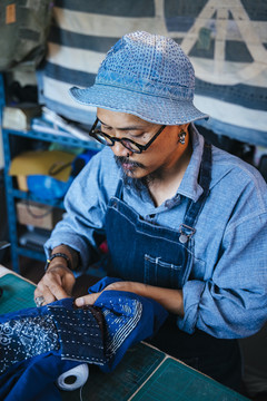 一个穿着蓝色衣服，戴着帽子和围裙的人边剪衣服边缝布料。