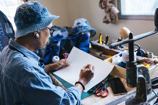 一个穿着蓝色衣服，戴着帽子，围裙的男人用铅笔在纸上画出自己衣服的图案，然后用智能手机在网上查看参考资