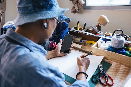 一个穿着蓝色衣服，戴着帽子，围裙的男人用铅笔在纸上画出自己衣服的图案，然后用智能手机在网上查看参考资