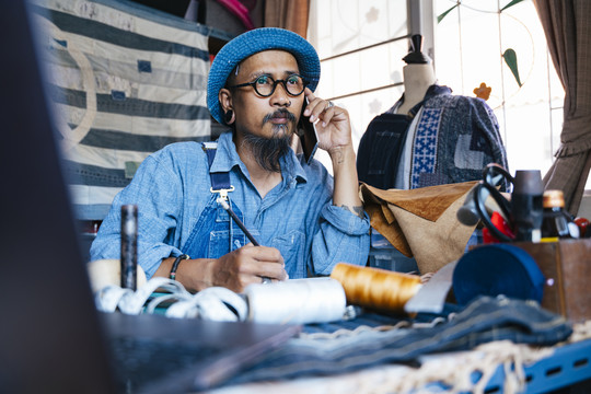 一个穿着蓝色衣服、帽子和围裙的男人用铅笔在纸上画出自己衣服的图案，然后用智能手机打电话给某人。