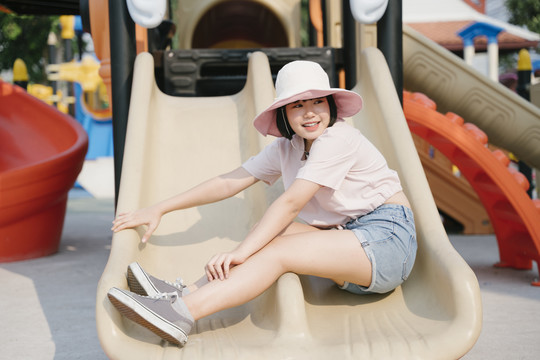 美丽的泰国亚裔女子戴着帽子，暑假喜欢在游乐场公园玩滑梯。