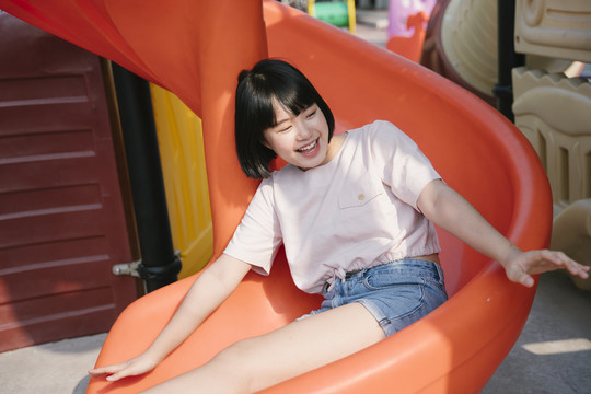 可爱的年轻亚泰女子鲍勃发型享受假期在游乐场公园坐在滑梯上。童年的快乐。坐在橙色的滑梯上。