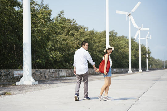 后景-快乐的亚泰年轻夫妇手牵手走在街上。风车背景。旅行者参观风车遗址。
