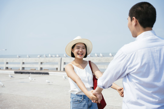 快乐的笑容亚泰女子穿着白衬衫戴着帽子牵着男友的手在海上大桥上旅行。