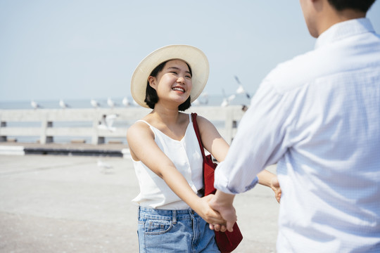 快乐的笑容亚泰女子穿着白衬衫戴着帽子牵着男友的手在海上大桥上旅行。
