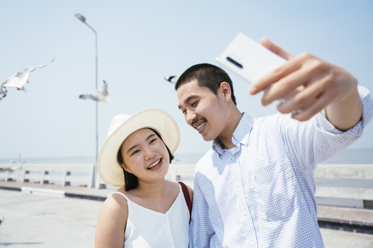 快乐的亚泰夫妇在海边大桥用智能手机拍照。海鸥在背景上飞翔。